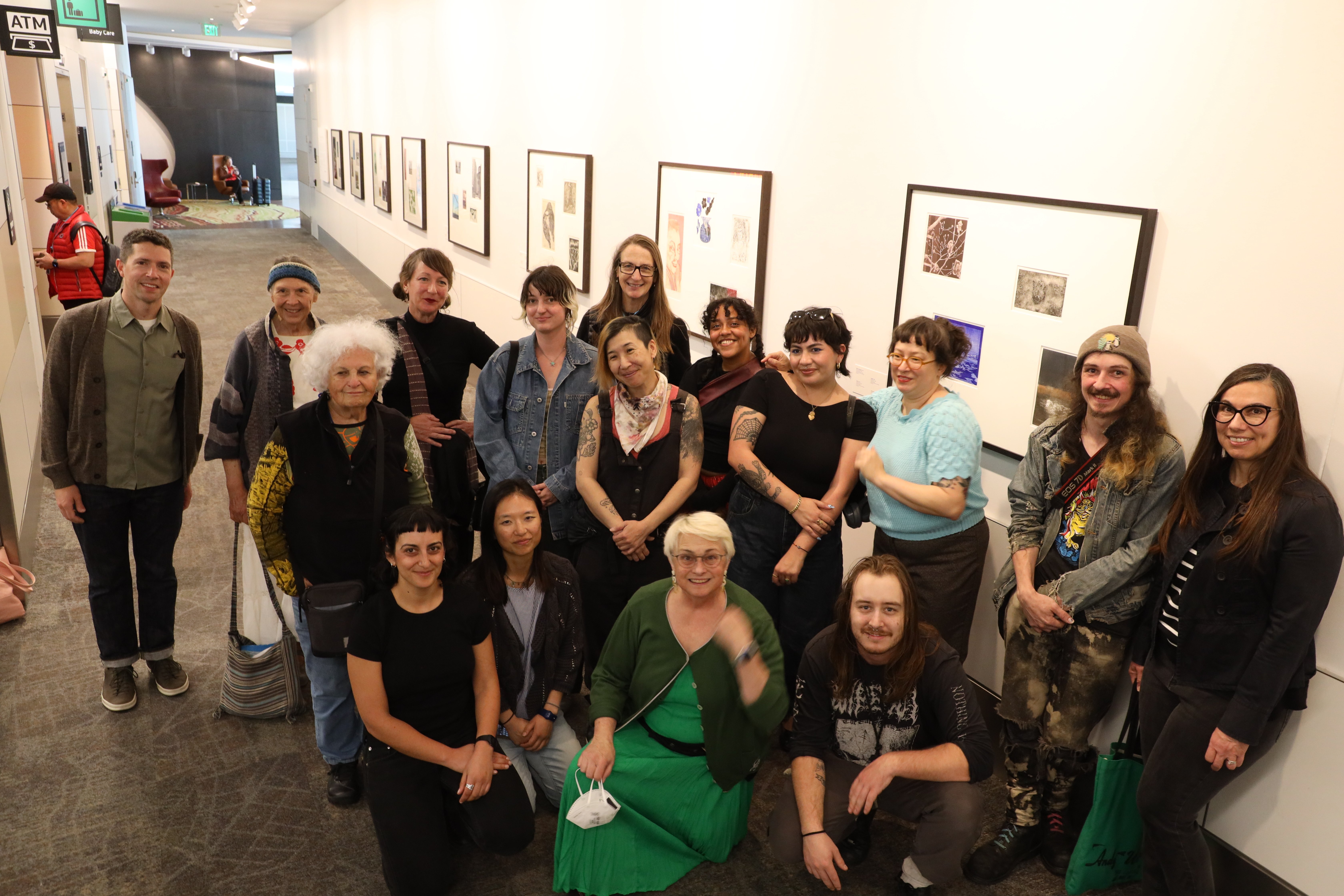 16 smiling people pose in front of a hallway filled with framed art prints. The people are Laney Professor Alex Echevarria plus the 15 Laney students with art prints featured in the exhibit at SFO. Photo by Laney photography professor Scot Tucker.