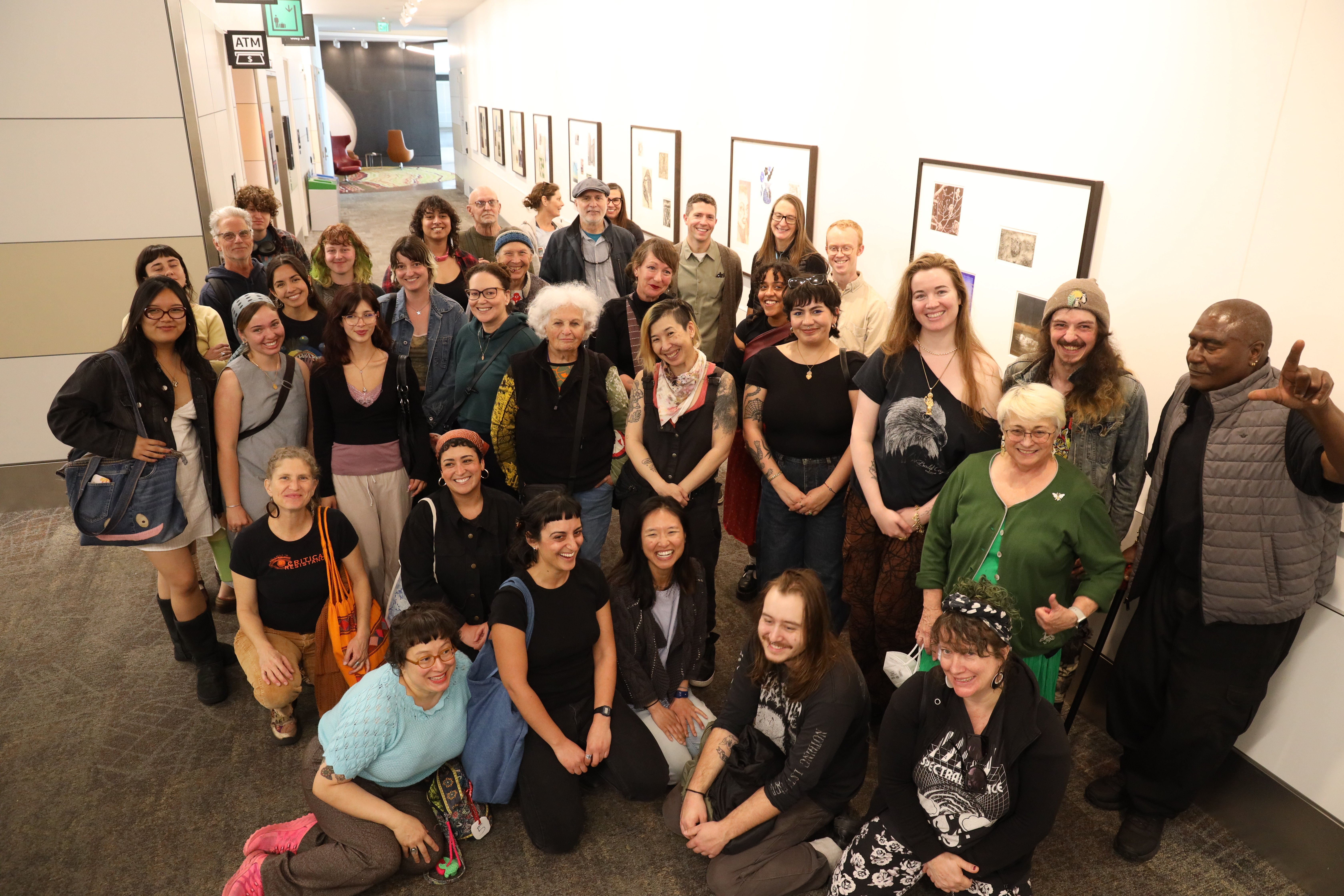 About 35 smiling people are standing in front of art framed on the wall of a long hallway. These are printmaking students of Professor Alex Echevarria visiting the Laney art exhibit at San Francisco International Airport. Photo taken by Laney photography professor Scot Tucker.