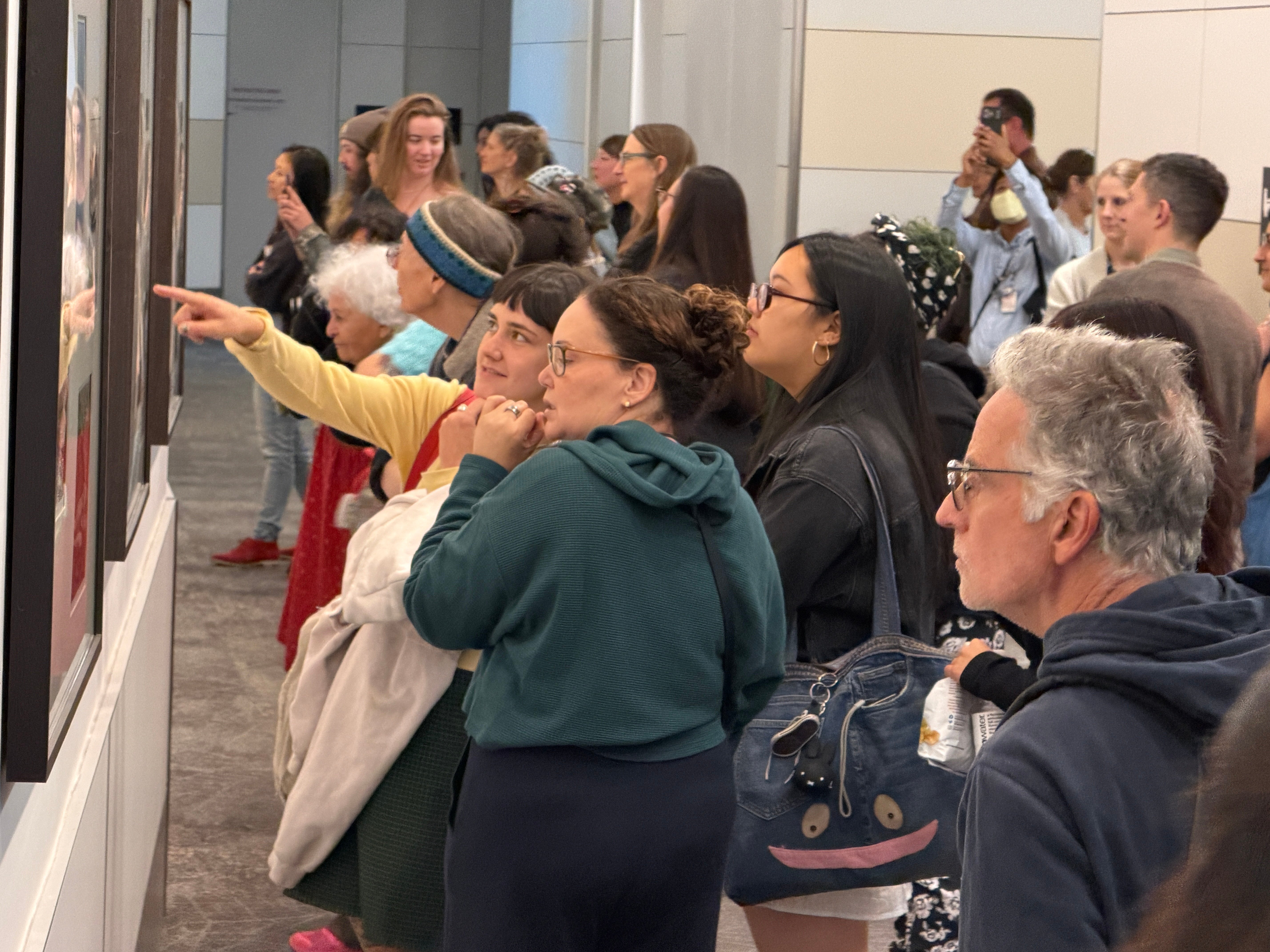 A group of approximately 20 people are standing close together looking at artwork on the wall of a long hallway. The people are Laney College printmaking students visiting an exhibit of Laney student art at San Francisco International Airport. Photo by Mark Johnson.