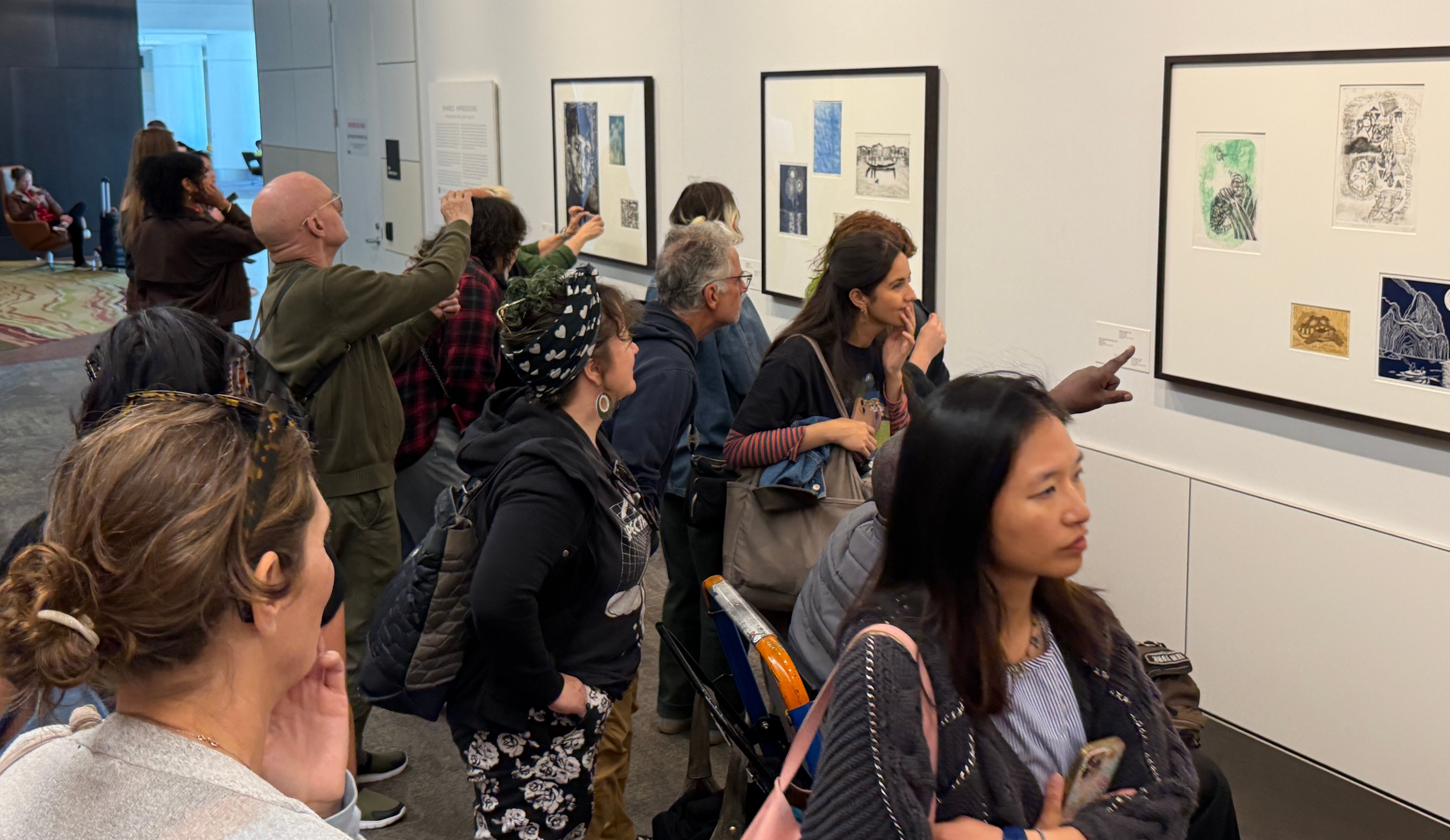 A diverse group of people are looking at framed artwork on a wall of a long hallway. The people are Laney College printmaking students enjoying an exhibit of Laney art on display at San Francisco International Airport. Photo by Mark Johnson.