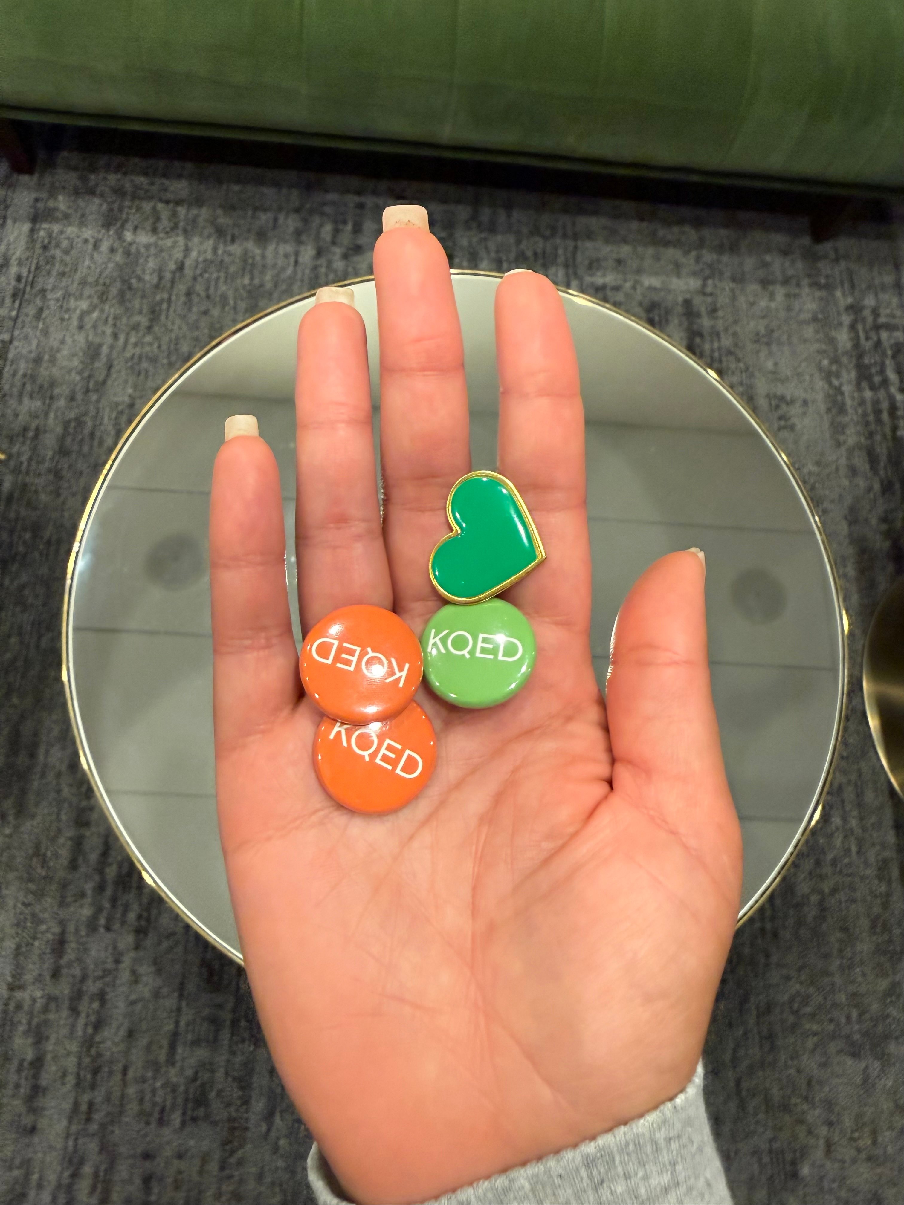 A hand is holding four pins, three round pins that say "KQED" (two of them orange, one green), and a green heart Laney Love pin.
