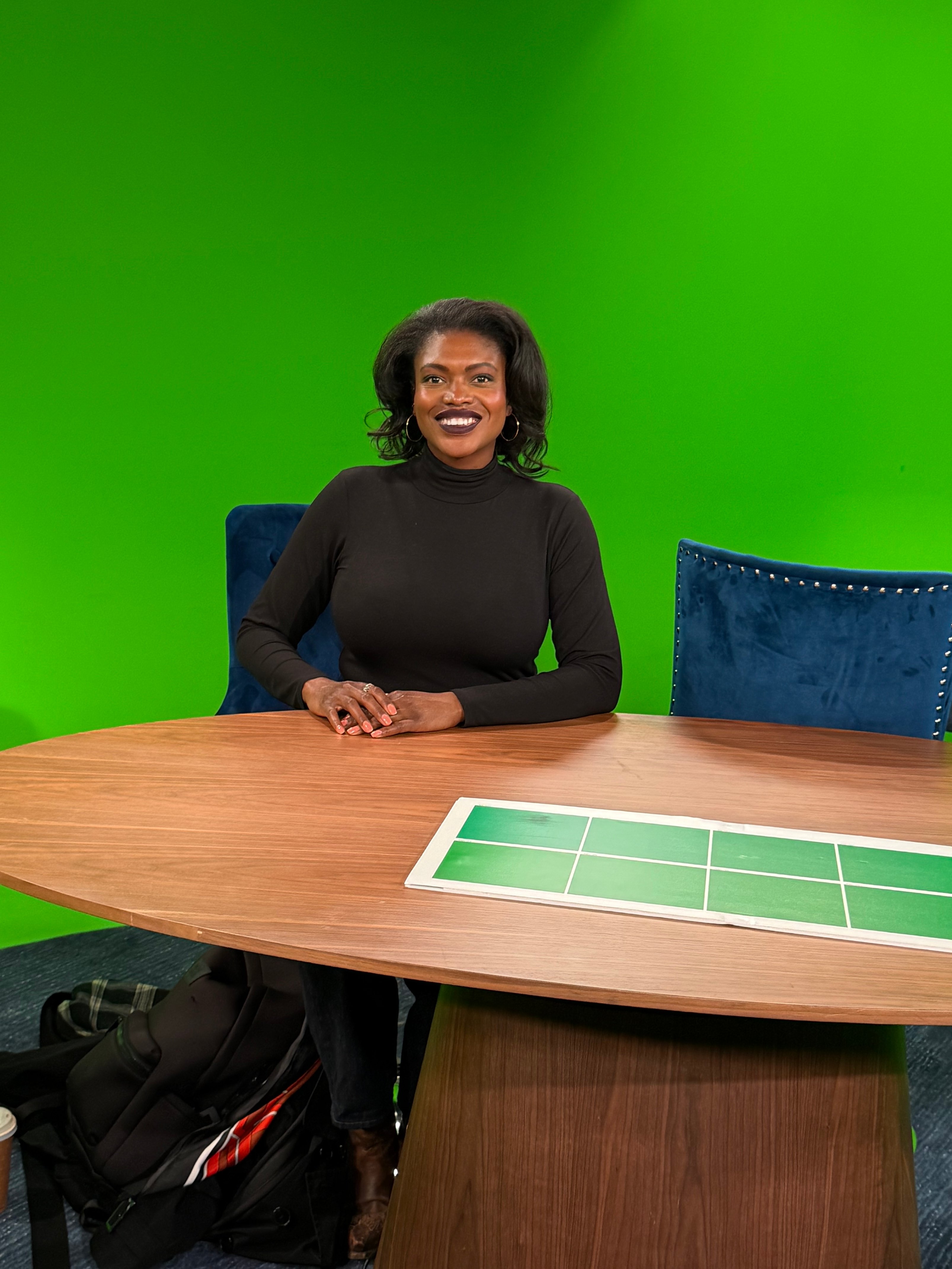 A smiling woman in a black sweater sits at a table in front of a green screen. The woman is Laney student Tamera Copes at the KQED studio in San Francisco.